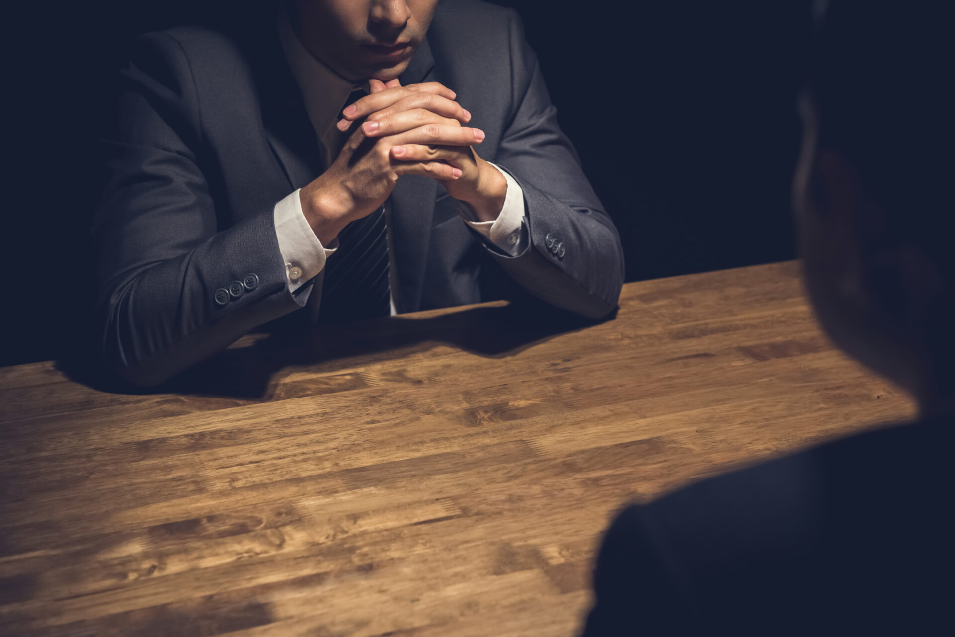 A sharply dressed man intently interrogating another male, both seated at a wooden table, engaged in a tense conversation.