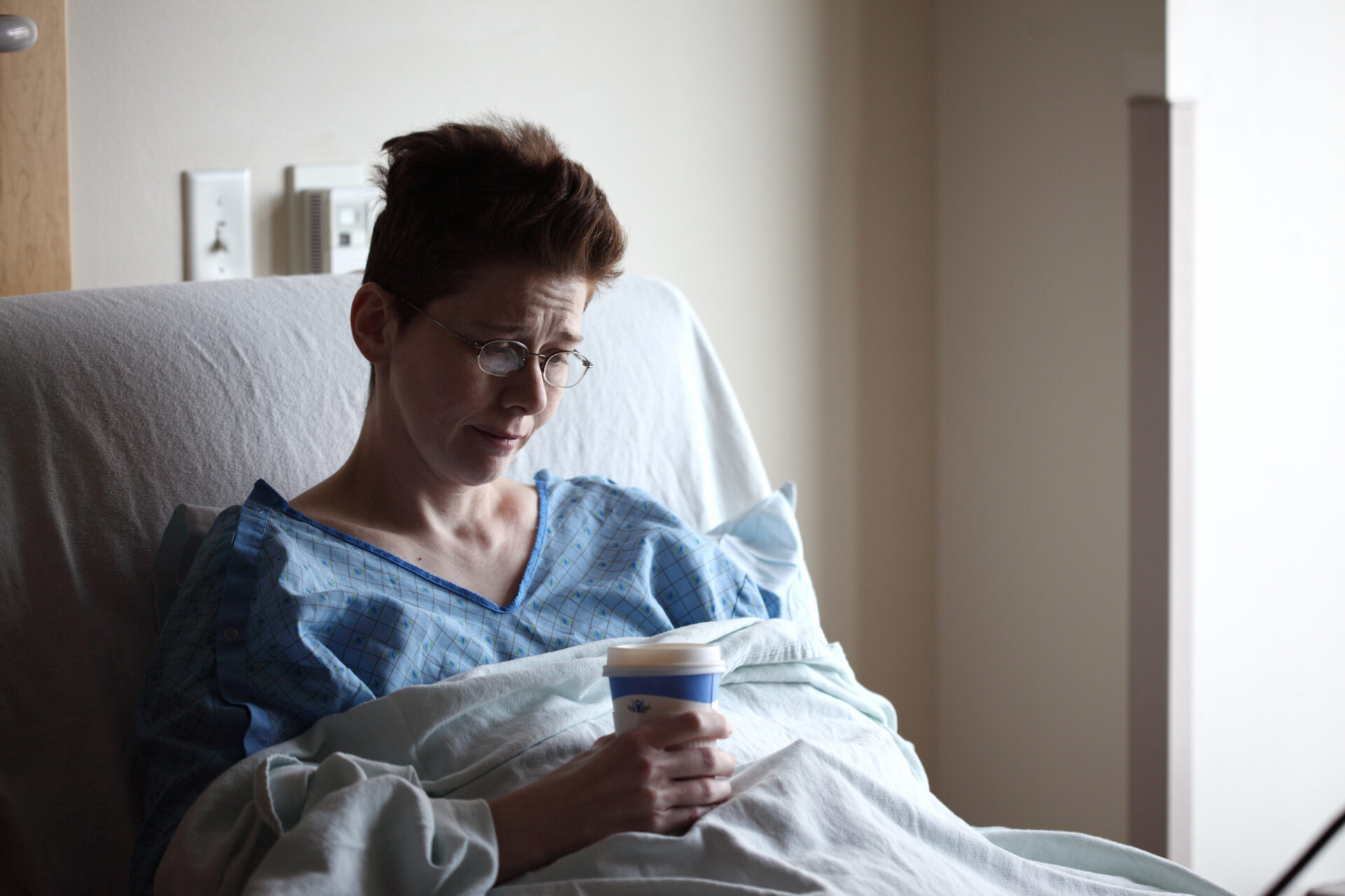 a women patient in hospital garments on a hospital bed with a hot drink in her hand